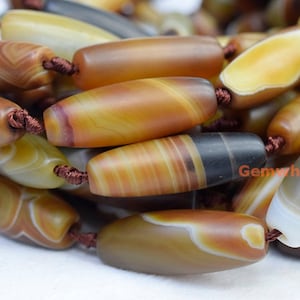 Può includere: Una collana di perline di agata marroni, gialle e bianche con un filo marrone. Le perline sono di forma ovale e hanno un motivo a strisce.