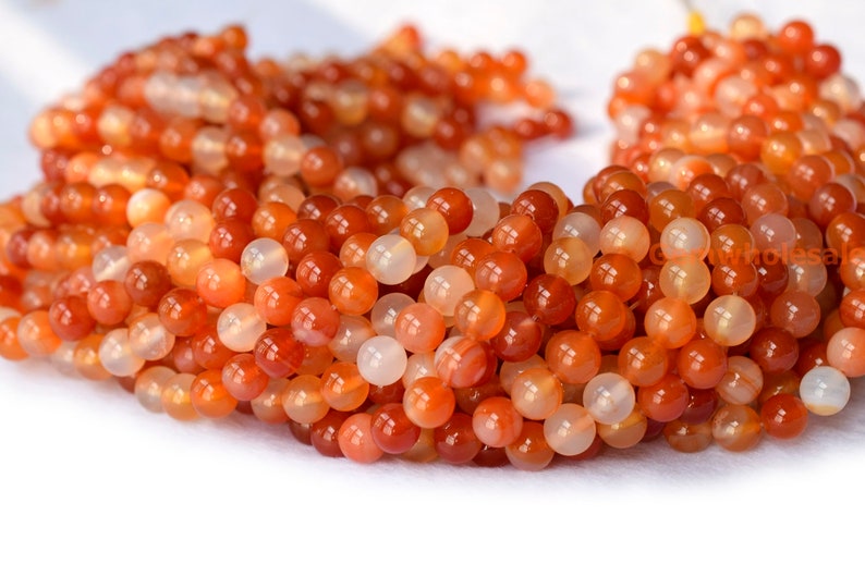 May include: A strand of orange carnelian beads. The beads are round and smooth, with a polished finish. They are a mix of light and dark orange shades.