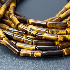 May include: A strand of tiger's eye beads, each bead is a long, cylindrical shape with a golden brown color and a dark brown stripe running through the center. The beads are arranged in a loose pile, showing the intricate patterns of the stone.