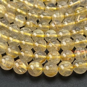 May include: A strand of round, polished rutilated quartz beads. Each translucent bead features golden, needle-like inclusions radiating throughout the stone. The beads are arranged in multiple rows, showcasing the natural variations in the golden rutile patterns.