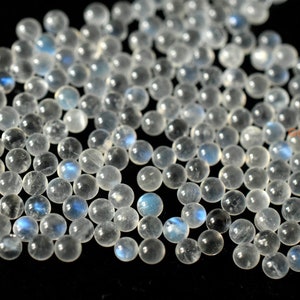 May include: A collection of round, white moonstone beads with a blue iridescent sheen. The beads are smooth and polished, and they are scattered on a black background.