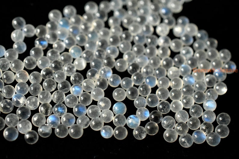 May include: A collection of round, white moonstone beads with a blue iridescent sheen. The beads are smooth and polished, and they are scattered on a black background.