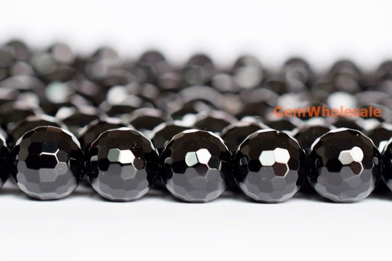 May include: Close-up of a strand of faceted black onyx beads. The beads are round with a multi-faceted surface, reflecting light. The beads are strung together, creating a dark, textured pattern. The background is white.