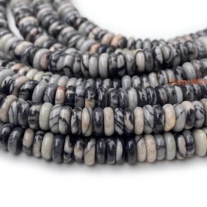 Puede incluir: Primer plano de hebras de cuentas de piedras preciosas grises y negras. Las cuentas son cilíndricas con una forma plana en forma de disco. Las cuentas tienen una apariencia veteada con una mezcla de colores grises, negros y crema. Las cuentas están ensartadas, creando un efecto en capas.