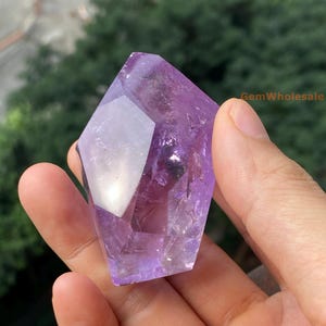 Puede incluir: Un gran cristal de amatista facetado de color morado oscuro. El cristal tiene forma de punta y una superficie lisa y pulida.