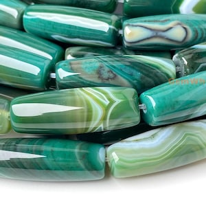 May include: Close-up of a collection of green agate beads. The beads are cylindrical with rounded ends and a polished, glossy finish. The beads have a mix of green and white swirling patterns.