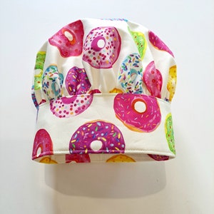 May include: White chef's hat with a donut print. The donuts are pink, green and yellow with colourful sprinkles.
