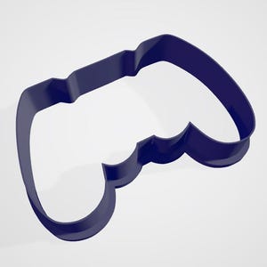 Może przedstawiać: Ciemnoniebieska foremka do ciastek w kształcie kontrolera do gier wideo. Foremka ma zaokrągloną górę i dół z uchwytem pośrodku. Projekt jest prosty i nowoczesny, idealny do tworzenia tematycznych ciasteczek.