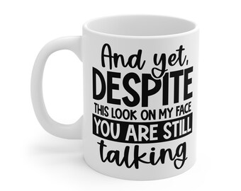 May include: White ceramic mug with a black text design. The text reads "And yet, DESPITE THIS LOOK ON MY FACE YOU ARE STILL talking". The mug has a curved handle.