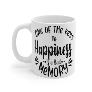 Op de afbeelding: Witte keramische koffiemok met een gebogen handvat. De mok heeft zwarte tekst die luidt: "One of the keys to Happiness is a bad memory."