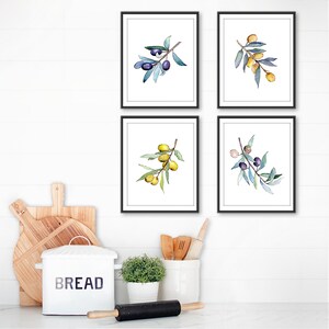 Op de afbeelding: Vier ingelijste aquarelprints van olijftakken met groene bladeren en zwarte olijven. De prints zitten in zwarte lijsten en worden op een witte muur getoond. Keukeninrichting met een brooddoos en deegroller op de voorgrond.