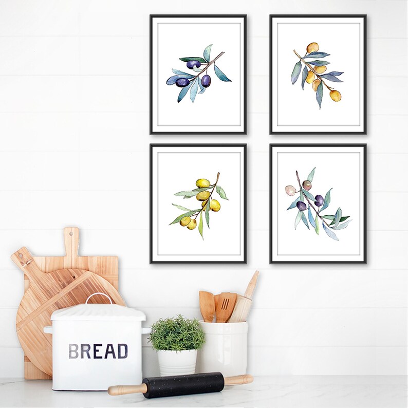 Op de afbeelding: Vier ingelijste aquarelprints van olijftakken met groene bladeren en zwarte olijven. De prints zitten in zwarte lijsten en worden op een witte muur getoond. Keukeninrichting met een brooddoos en deegroller op de voorgrond.