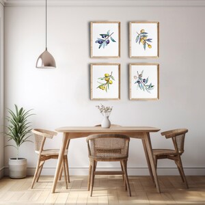 Op de afbeelding: Vier ingelijste aquarelprints van olijftakken met groene bladeren en paarse of gele olijven hangen aan een witte muur. Een houten eettafel en stoelen staan voor de muur, met een potplant en een vaas met bloemen.