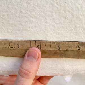 May include: A close-up of a wooden ruler with black markings, showing measurements from 1 to 6 inches. The ruler is resting on a white textured surface.