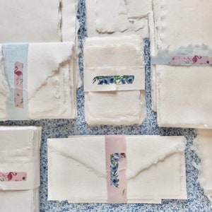 May include: A stack of white handmade paper sheets with decorative washi tape accents. The tape features floral and flamingo designs.