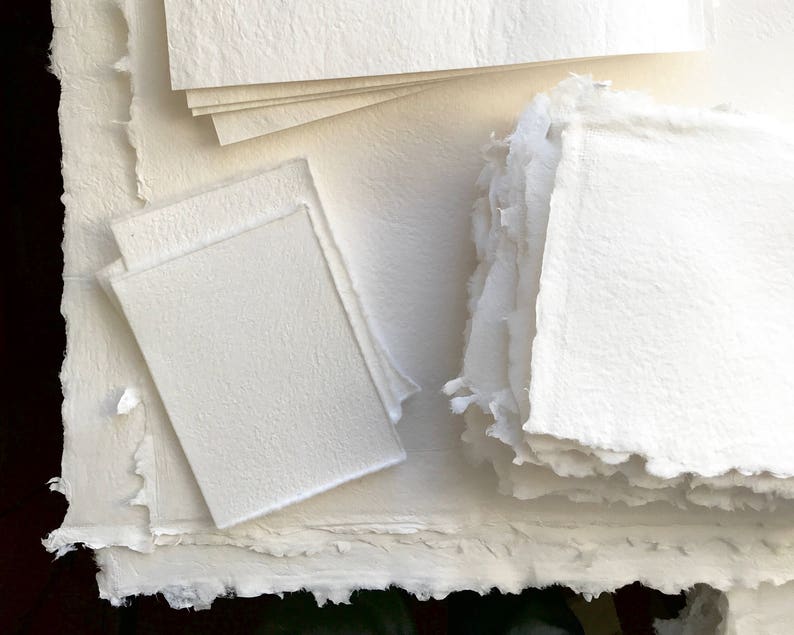 May include: A stack of white handmade paper sheets with a textured surface. The paper is arranged in a variety of sizes and shapes.