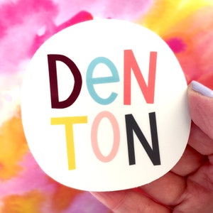 May include: A white circle sticker with the word 'DENTON' written in colorful letters. The letters are in different colors: maroon, light blue, pink, yellow, pink, and black.