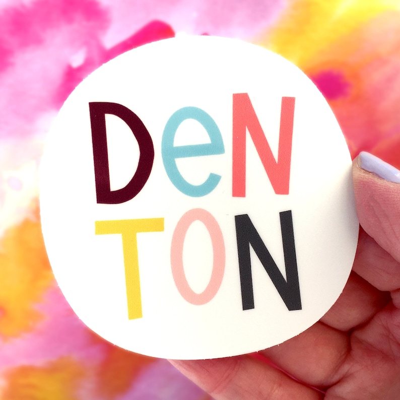 May include: A white circle sticker with the word 'DENTON' written in colorful letters. The letters are in different colors: maroon, light blue, pink, yellow, pink, and black.