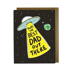 May include: A rectangular greeting card with a black background featuring white stars and a green planet. A light blue UFO emits a bright yellow beam of light containing the black text: YOU'RE THE BEST DAD OUT THERE. A brown envelope is visible behind.