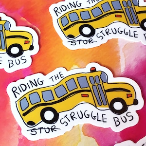 May include: A yellow school bus sticker with the text "Riding the Struggle Bus" in black. The bus is tilted on its side, with the wheels facing upwards. The sticker is on a pink and orange watercolor background.