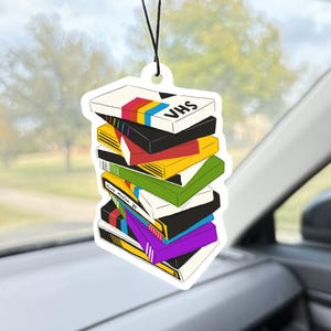 May include: A stack of colorful VHS tape air fresheners hanging from a black string. The air fresheners are in the shape of VHS tapes, with the word "VHS" printed on the top tape. The tapes are various colors, including black, yellow, red, and purple.