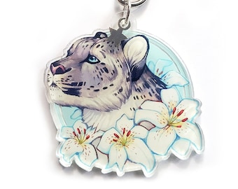 Puede incluir: Un leopardo de las nieves blanco con ojos azules está rodeado de lirios blancos sobre un fondo azul claro. La imagen está en un llavero con un anillo de metal plateado.