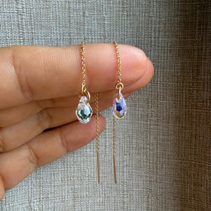 Puede incluir: Un par de pendientes de hilo dorados. Cada pendiente presenta un colgante de cristal facetado e iridiscente y una delicada cadena. Los pendientes se muestran sobre un fondo neutro, mostrando su elegante diseño.