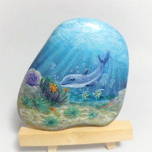 May include: Hand-painted rock art depicting a dolphin swimming in a blue ocean with coral reefs, fish, and jellyfish. The rock is displayed on a wooden easel.