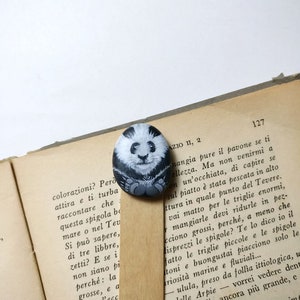 May include: A hand-painted rock bookmark featuring a black and white panda bear with a cute expression. The bookmark is resting on a page of a book with Italian text.