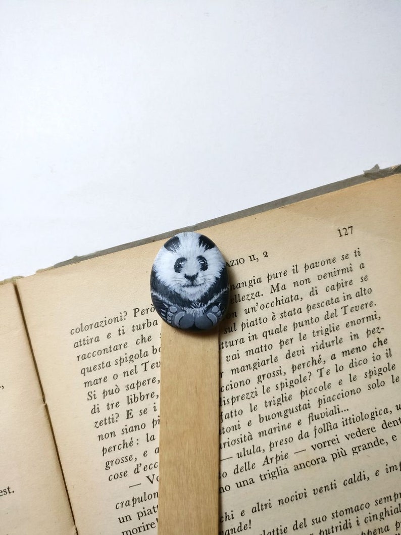 May include: A hand-painted rock bookmark featuring a black and white panda bear with a cute expression. The bookmark is resting on a page of a book with Italian text.