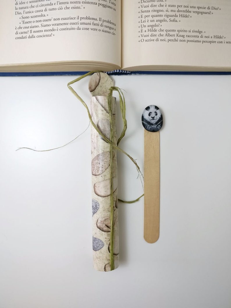 May include: A close-up of a rolled up piece of paper with a birch bark pattern. The paper is tied with a green string. A wooden bookmark with a panda bear illustration is to the right of the paper.