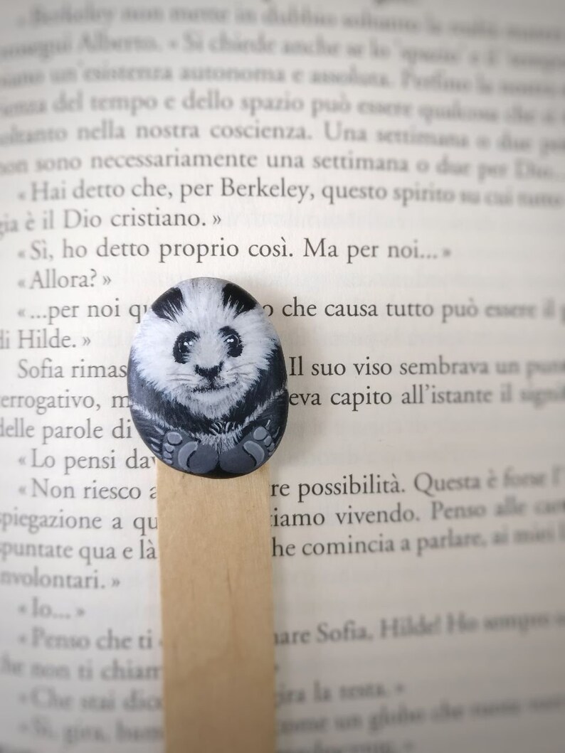 May include: A wooden bookmark with a hand-painted black and white panda bear design. The panda is sitting with its paws in front of it. The bookmark is resting on a page of a book.