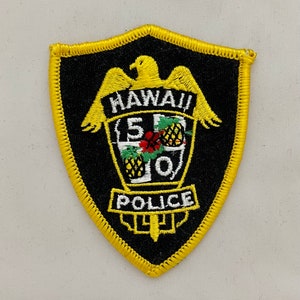 May include: Embroidered black patch with a yellow border and a shield shape. The patch features a yellow eagle above the word "HAWAII" and the text "POLICE". The center design includes pineapples and the numbers "5-0".