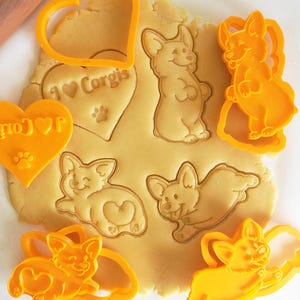 May include: A set of orange plastic cookie cutters pressed into rolled dough, featuring designs of a heart with the text I Love Corgis and a paw print, a standing Corgi, a lying Corgi, and a jumping Corgi, with the text CrimsonMane Creations below.