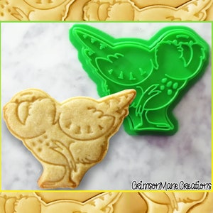 May include: A bird-shaped biscuit next to a green plastic cookie cutter. The biscuit is light brown with detailed impressions. The cookie cutter is bright green. The background is white with a marble effect. The text "Crimson Mane Creations" is visible.