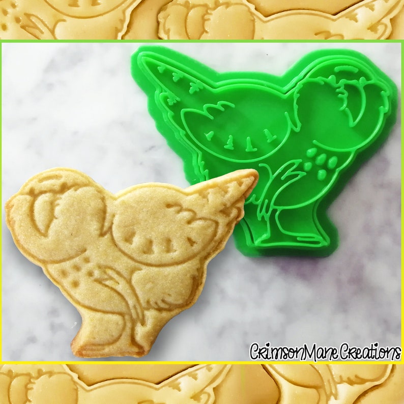 May include: A baked cookie in the shape of a bird next to a green plastic cookie cutter. The cookie is light brown with detailed impressions. The cookie cutter is green and has the same bird shape. The text "Crimson Mane Creations" is in the bottom right corner.