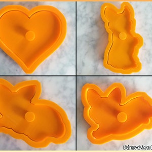 May include: A set of four orange plastic cookie cutters featuring a heart shape and three distinct corgi dog poses, including one standing, one sitting, and one lying down, with the text CrimsonMane Creations printed in the bottom right corner.