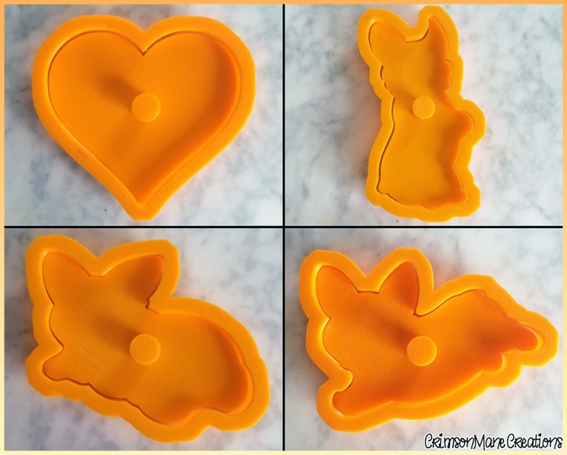 May include: A set of four orange plastic cookie cutters featuring a heart shape and three distinct corgi dog poses, including one standing, one sitting, and one lying down, with the text CrimsonMane Creations printed in the bottom right corner.