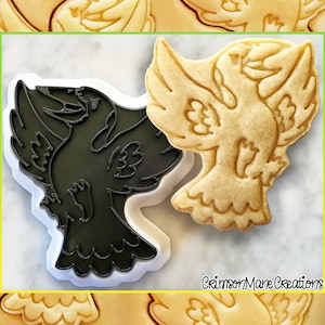 Può includere: Un tagliapasta nero a forma di uccello con le ali spiegate. Accanto al tagliapasta c'è un biscotto a forma di uccello con le ali spiegate. Il biscotto è di colore marrone chiaro e presenta un design dettagliato. Il testo "CrimsonMare Creations" è visibile nell'angolo in basso a destra dell'immagine.