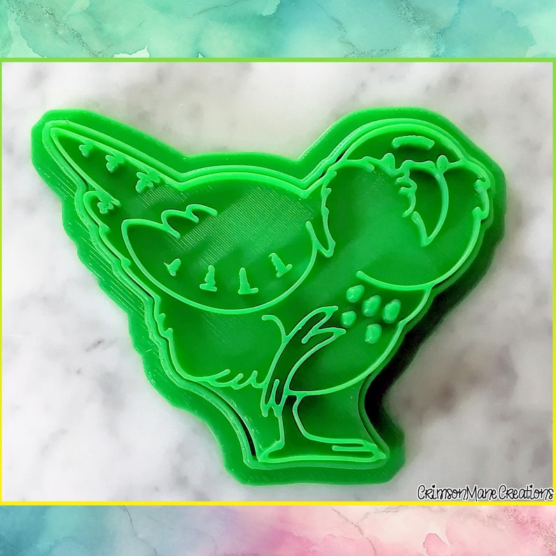 May include: A bright green bird-shaped cookie cutter. The cutter is made of plastic and has a detailed outline of a bird with a wing, tail, and eye. The cutter is designed to cut out bird-shaped cookies or other treats. The background is a marble-like surface.