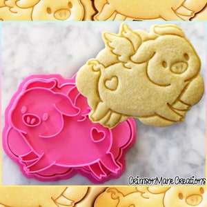 May include: Pink plastic cookie cutter shaped like a pig with wings and a heart on its side. A cookie in the shape of a pig with wings is on top of the cutter.