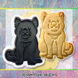 May include: A black plastic dog-shaped cookie cutter next to a baked cookie in the same shape. The cookie is golden brown. The cookie cutter has a white border. The text "Crimson Mane Creations" is at the bottom.