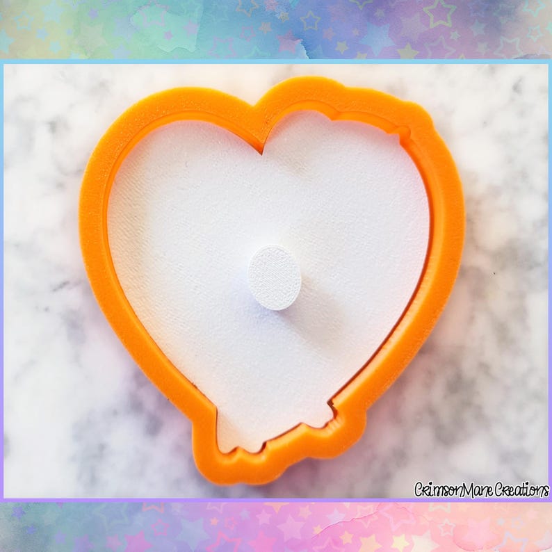 May include: An orange heart-shaped cookie cutter with a white center and a small white cylinder in the middle. The cutter has a smooth, rounded edge and is designed for baking. The background is a soft, colorful gradient.