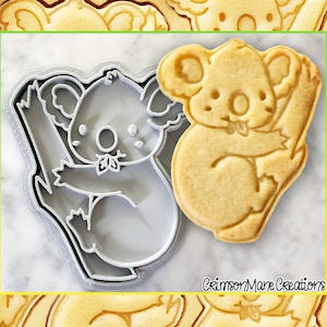 May include: A grey plastic koala cookie cutter and a light brown sugar cookie in the shape of a koala. The cookie cutter is on the left and the cookie is on the right. The cookie cutter is a detailed outline of a koala holding onto a tree branch. The cookie is a solid shape of a koala holding onto a tree branch. The cookie cutter and cookie are on a white surface. The text "CrimsonMare Creations" is in the bottom right corner of the image.