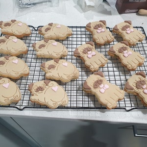 May include: A cooling rack with 12 decorated sugar cookies shaped like pugs. The cookies are brown with pink accents and some have a pink bow.