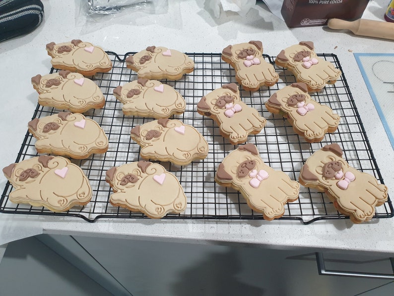 May include: A cooling rack with 12 decorated sugar cookies shaped like pugs. The cookies are brown with pink accents and some have a pink bow.