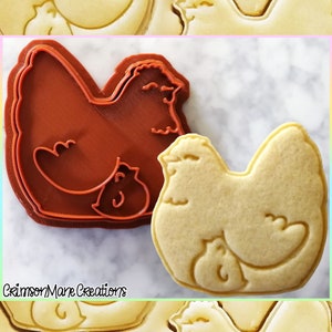 May include: A brown plastic cookie cutter shaped like a hen with a chick under her wing. Next to it is a baked sugar cookie in the same shape, but in a light yellow color.