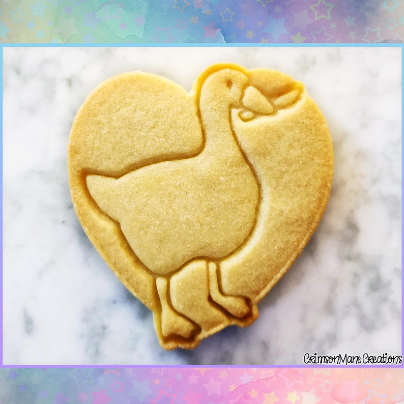 May include: A heart-shaped cookie with a goose design. The cookie is light brown with a detailed outline of a goose inside the heart shape. The cookie is on a white and gray marble surface.