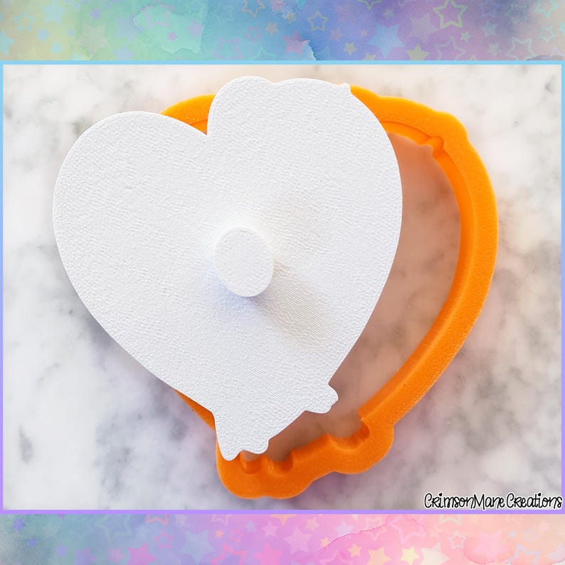 May include: A white heart-shaped cookie cutter with a small, round, white accent, set inside an orange heart-shaped frame. The cookie cutter is on a light gray and white marble surface.
