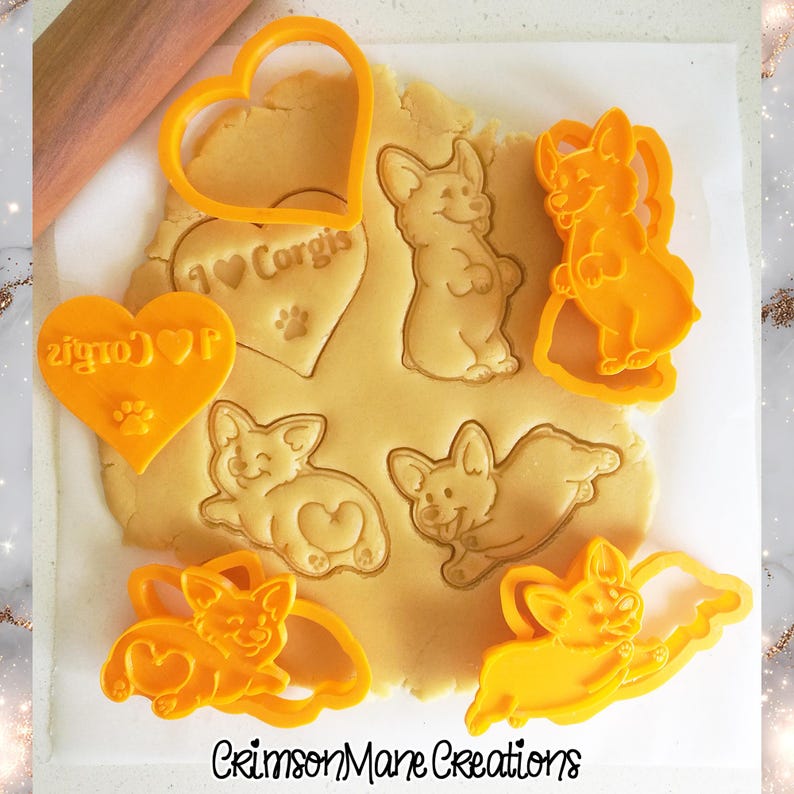 May include: A set of orange plastic cookie cutters pressed into rolled dough, featuring designs of a heart with the text I Love Corgis and a paw print, a standing Corgi, a lying Corgi, and a jumping Corgi, with the text CrimsonMane Creations below.